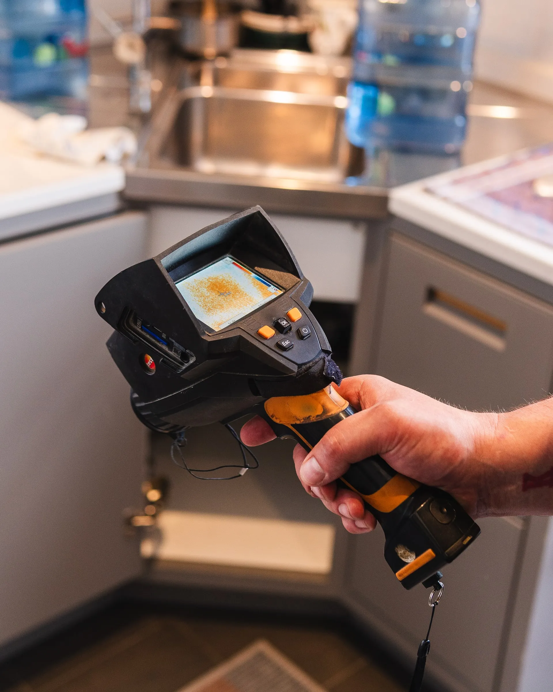 Close-up of a handheld thermal camera held over a floor, the screen showing a warm patch