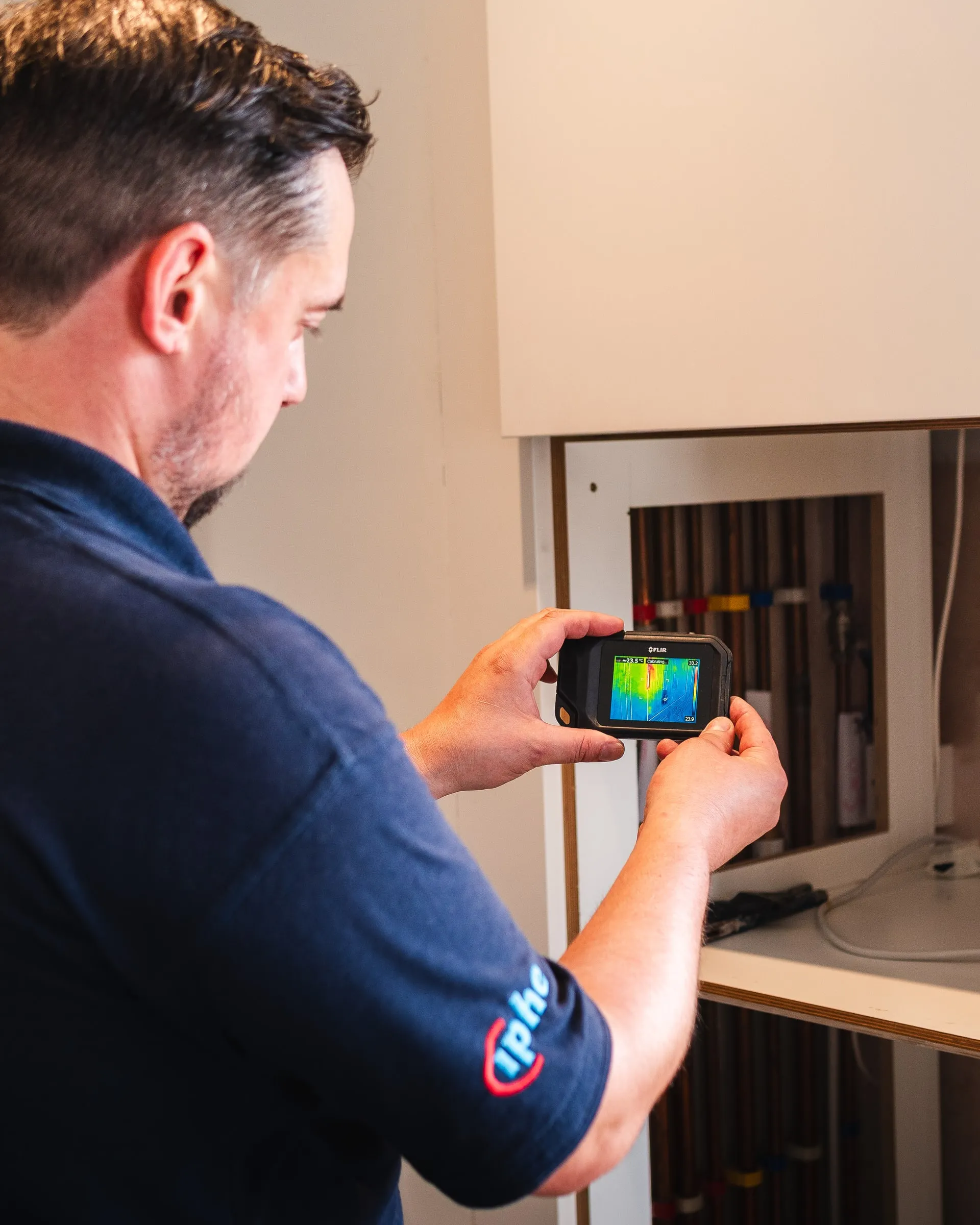 Engineer checking pipework inside an open cupboard with a thermal camera, the screen showing the warm pipe runs
